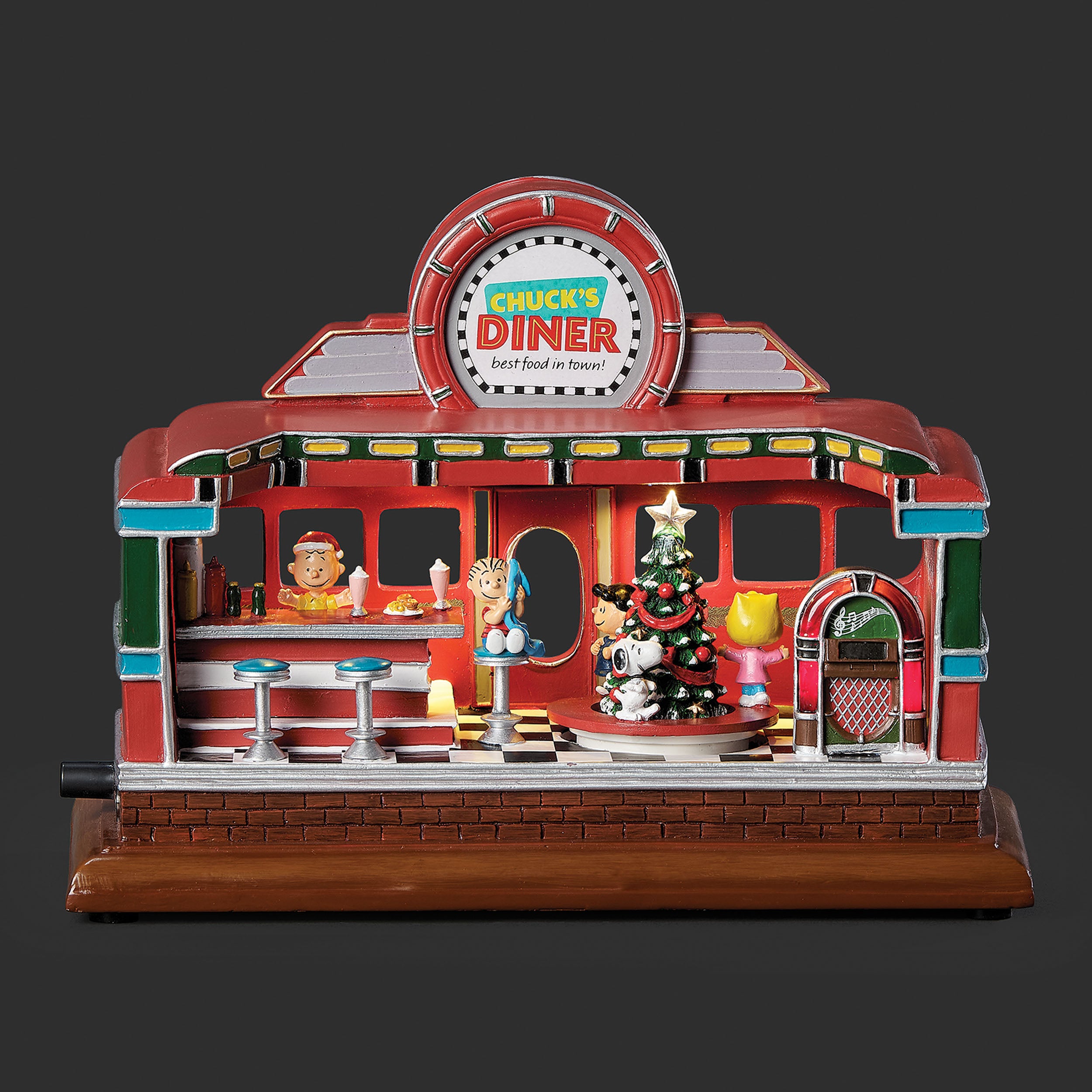 A decorative miniature toy diner set on a black background. The red and brown toy building features a circular sign reading 'Chuck's Diner' at the top, a counter, peanut characters, a green Christmas tree, and a jukebox on a brown base.