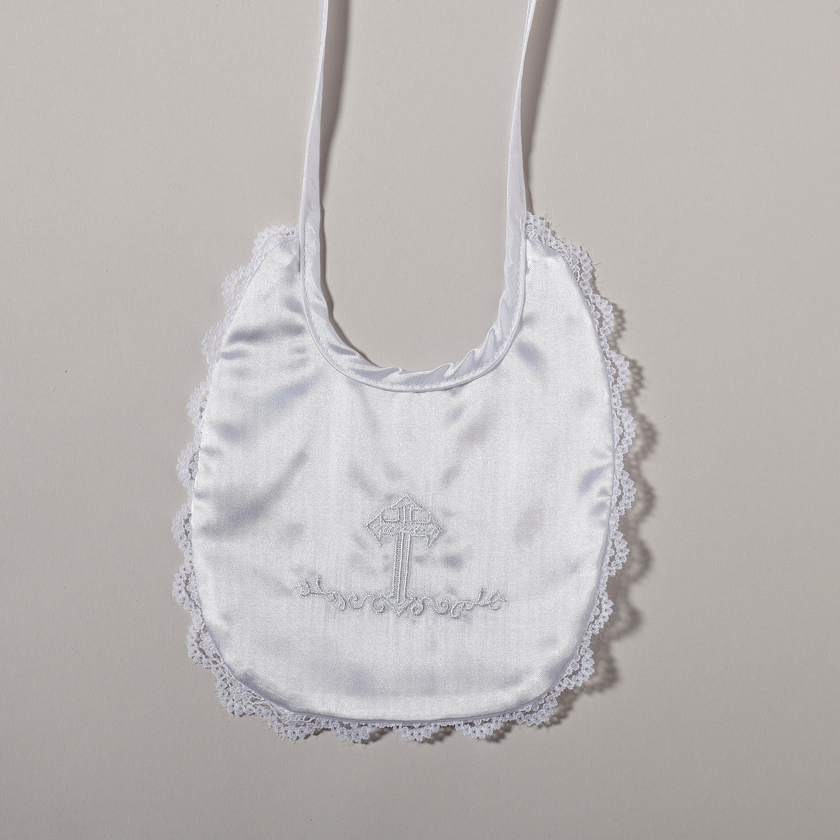 Front view of white baptism bib showing the embroidered cross, rounded shape, lace-trimmed edge, and ribbon ties