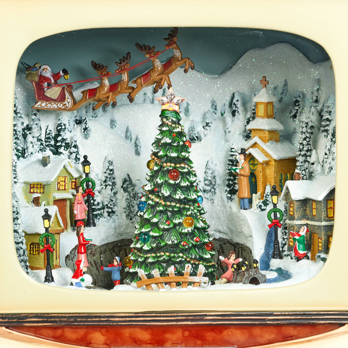 A festive Christmas diorama scene displayed inside a vintage television screen or television-shaped decorative frame. The scene features a decorated Christmas tree in the center, snow-covered houses, a church, street lamps, Santa Claus flying in a sleigh with reindeer, and small holiday figures, all set against a snowy mountain backdrop.