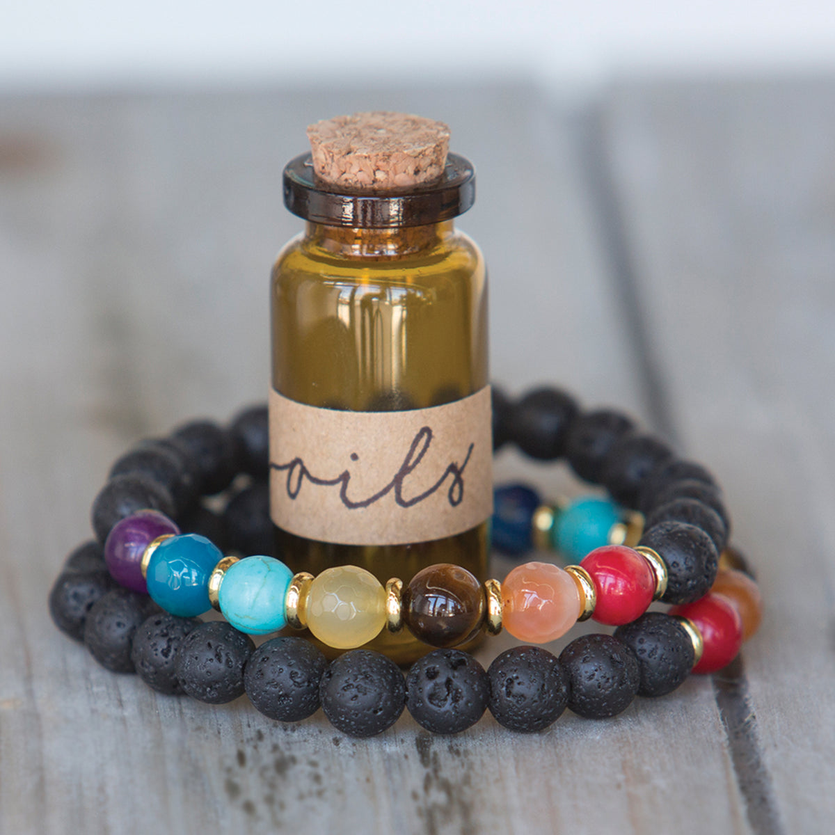 A small amber glass bottle with a cork stopper and a kraft paper label reading 'oils', styled in front of two beaded bracelets on a rustic gray wooden surface. One bracelet is made of black lava stones, and the other features multicolored gemstone beads separated by gold spacer rings.