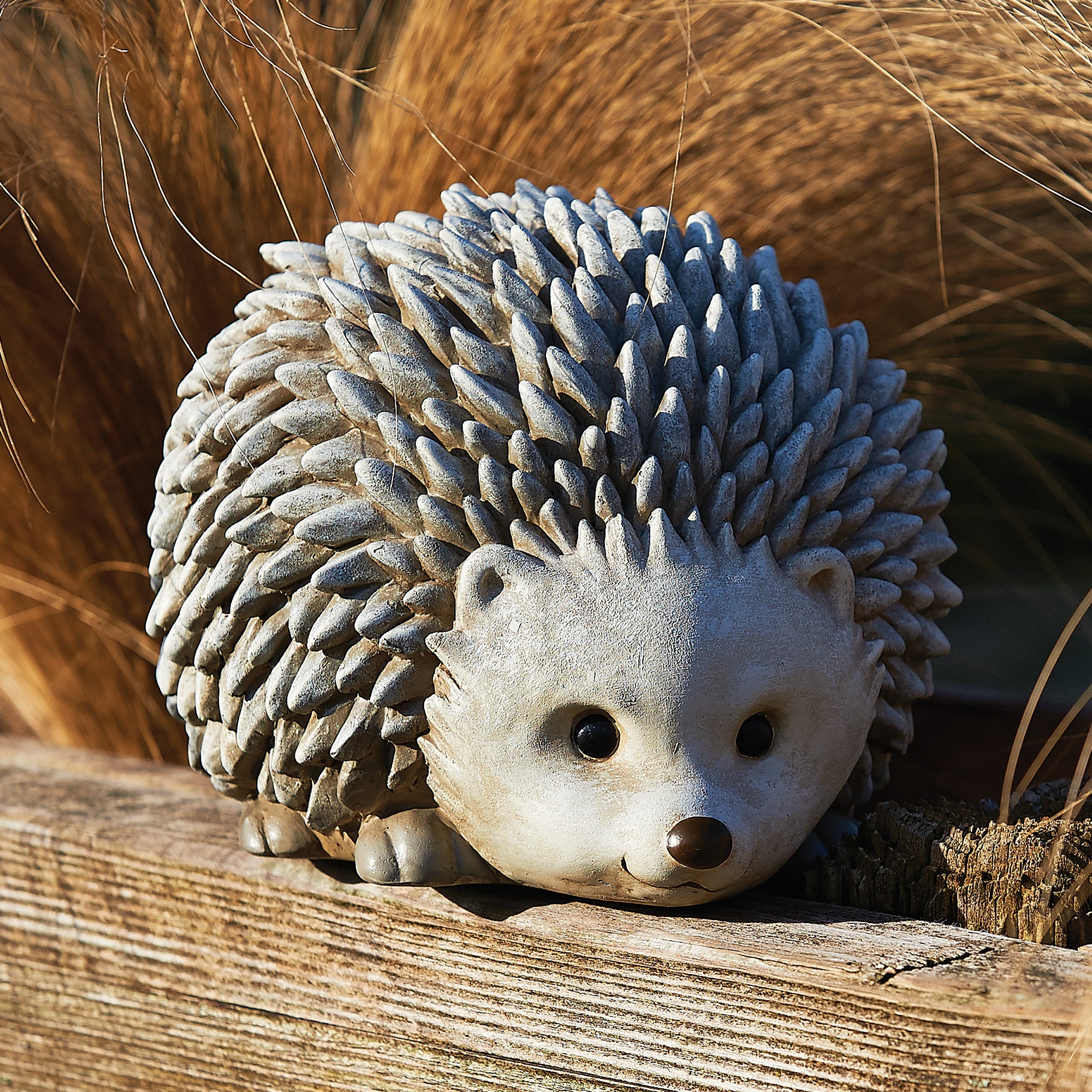 A small hedgehog figurine with gray spines and a cream-colored face, resting on a weathered wooden surface with dry grass in the background.