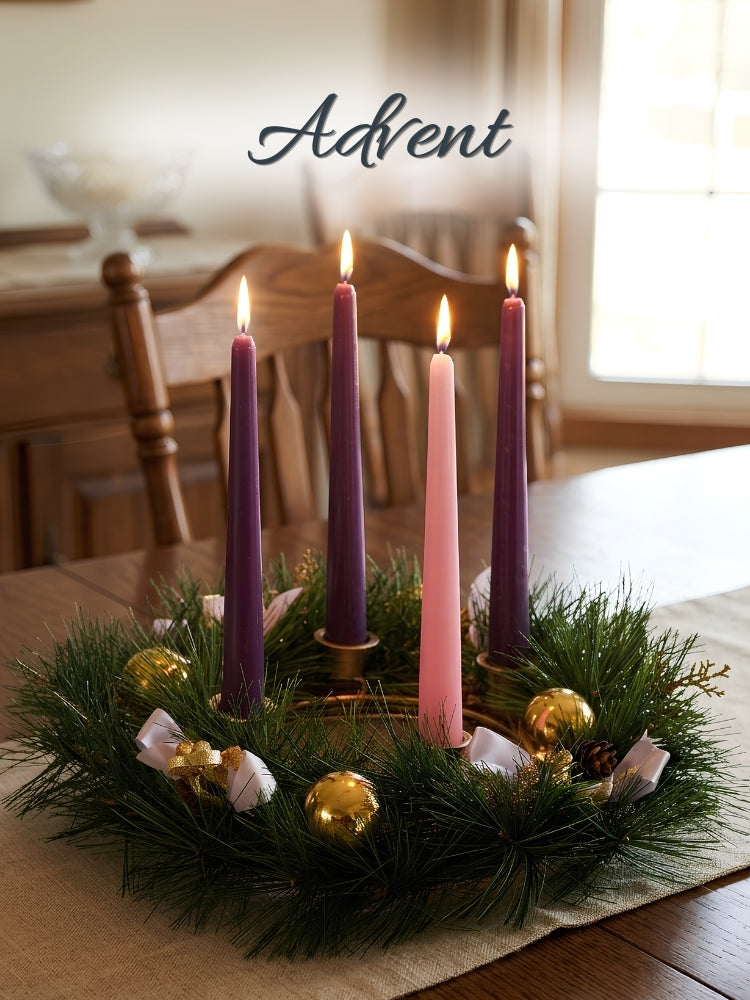 A festive Advent wreath centerpiece on a wooden table, featuring four taper candles in purple and pink, lit flames, green pine branches, gold ornaments, and white ribbons. The word 'Advent' is written in script at the top.