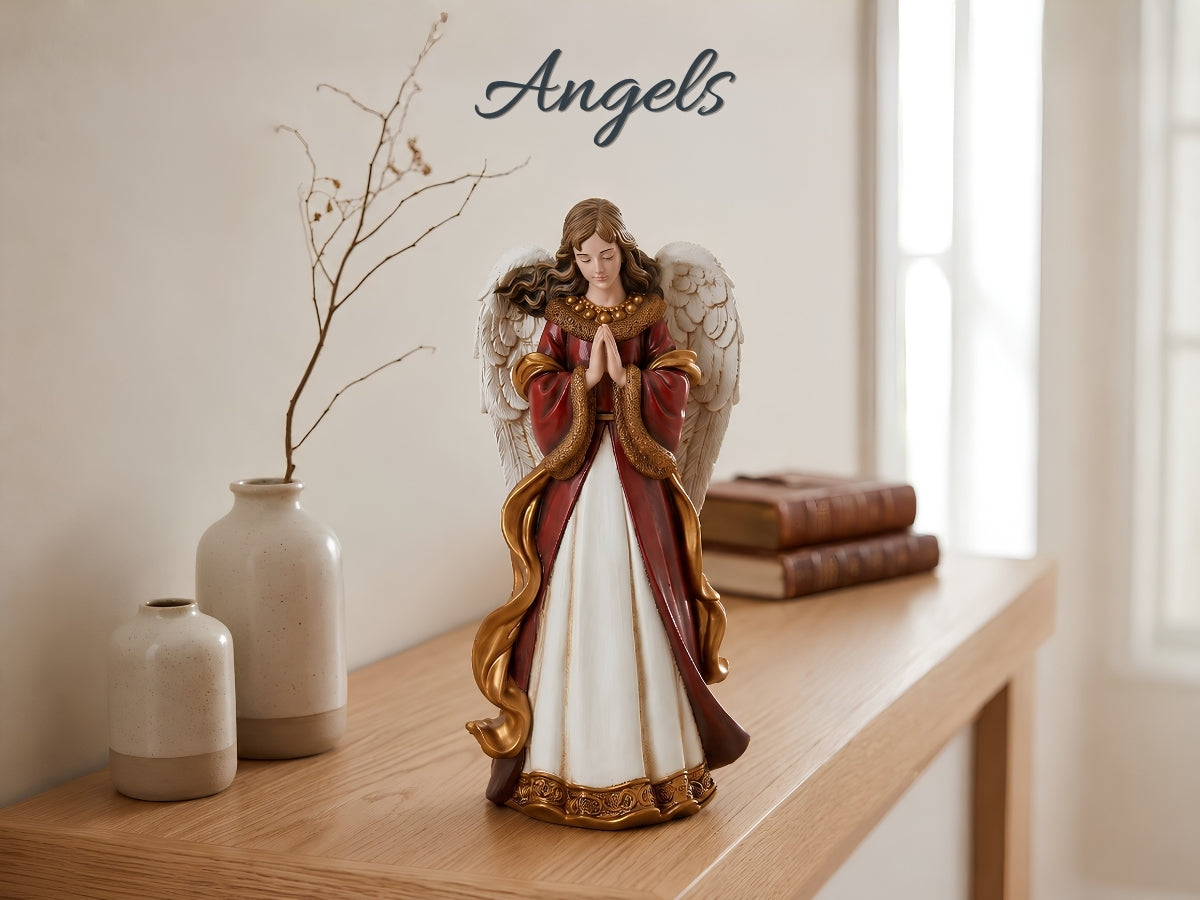 A praying angel figurine with gold wings and a red-and-white robe stands on a wooden console table in a softly lit room. Decorative vases, a branch, stacked books, and the word 'Angels' appear in the background.