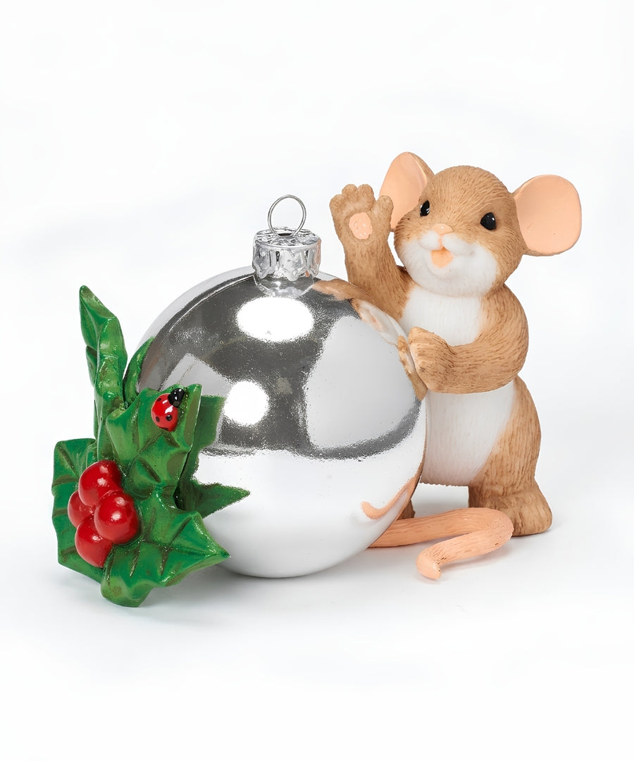 A Christmas ornament featuring a small brown and white mouse standing beside a shiny silver bauble, with green holly leaves and red berries on the left, photographed on a white background.