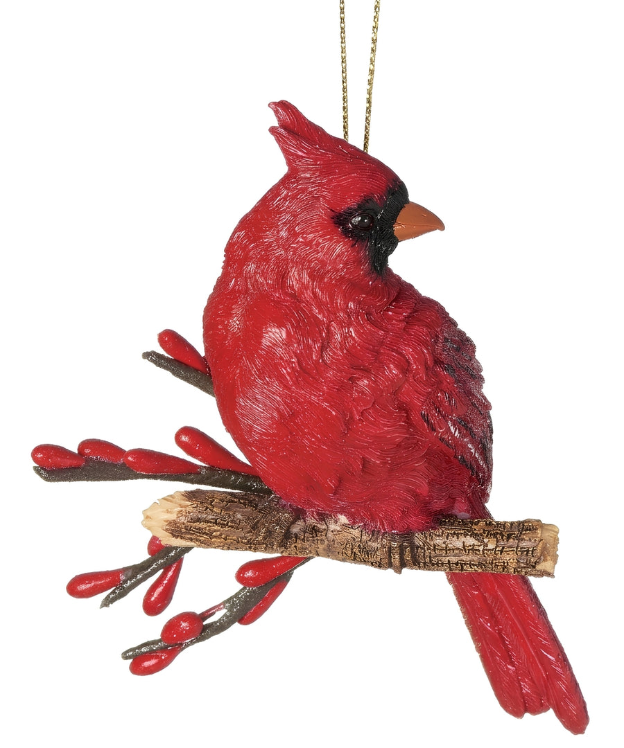 A red cardinal bird ornament perched on a small branch with red berries, hanging from a gold string against a plain white background.