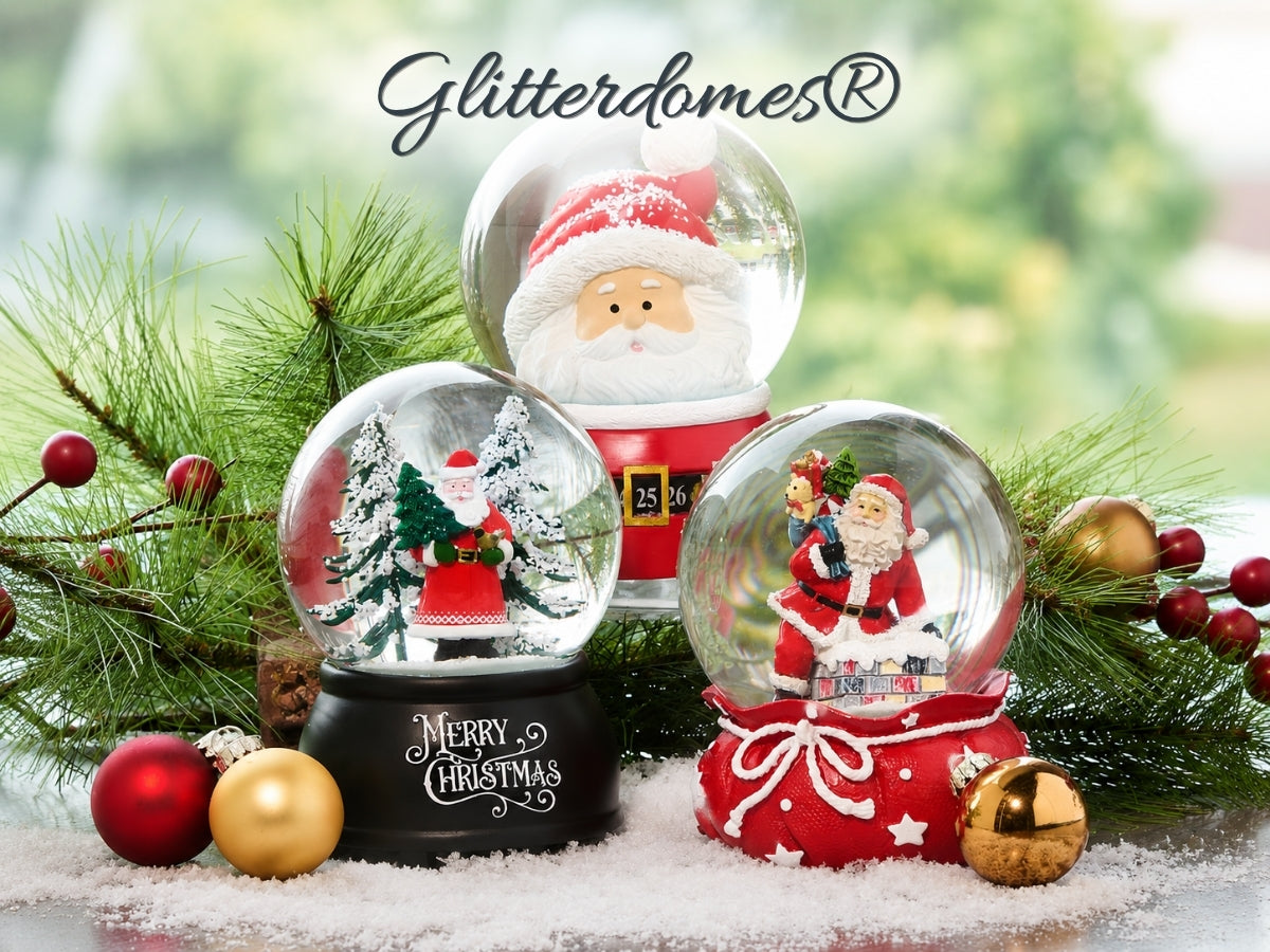 Three Glitterdomes Christmas snow globes displayed together with festive green pine branches, red and gold ornaments, and a blurred outdoor background. The globes feature Santa Claus figures and miniature holiday scenes, with the brand name Glitterdomes written at the top.