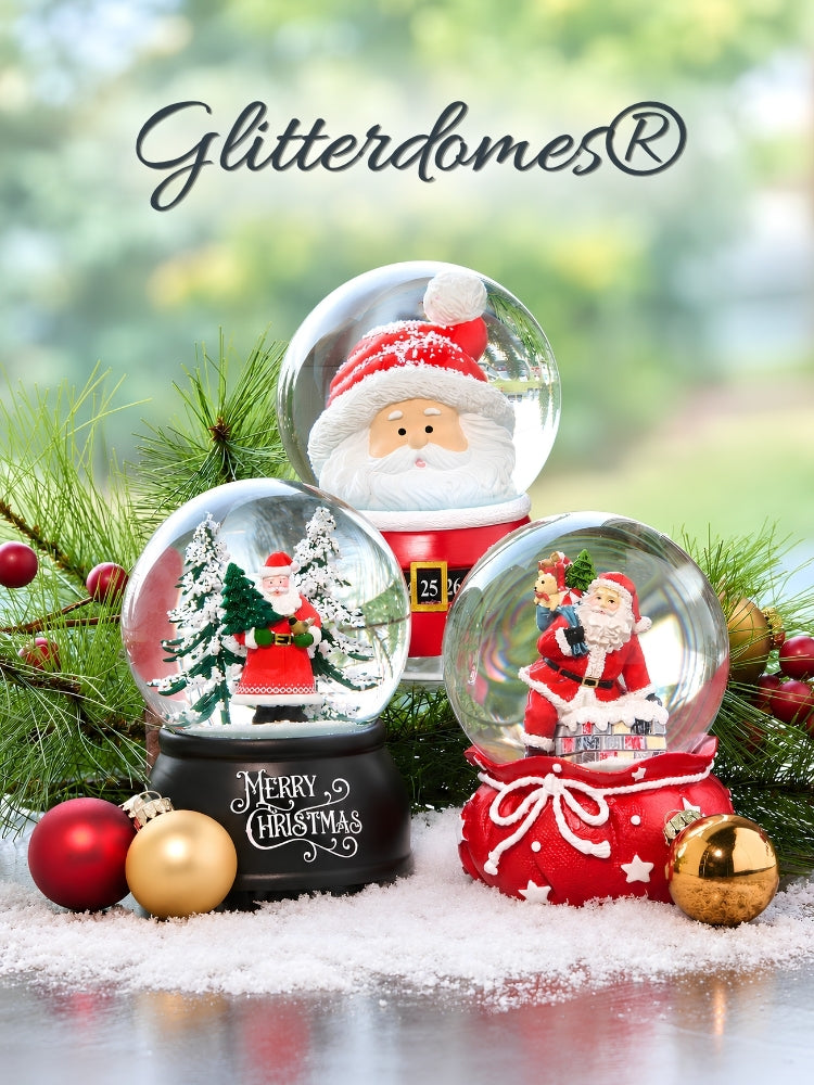 Three Christmas snow globes with Santa Claus figures and festive decorations, arranged on a reflective surface with pine branches, red and gold ornaments, and a blurred outdoor background. The word 'Glitterdomes' appears at the top.