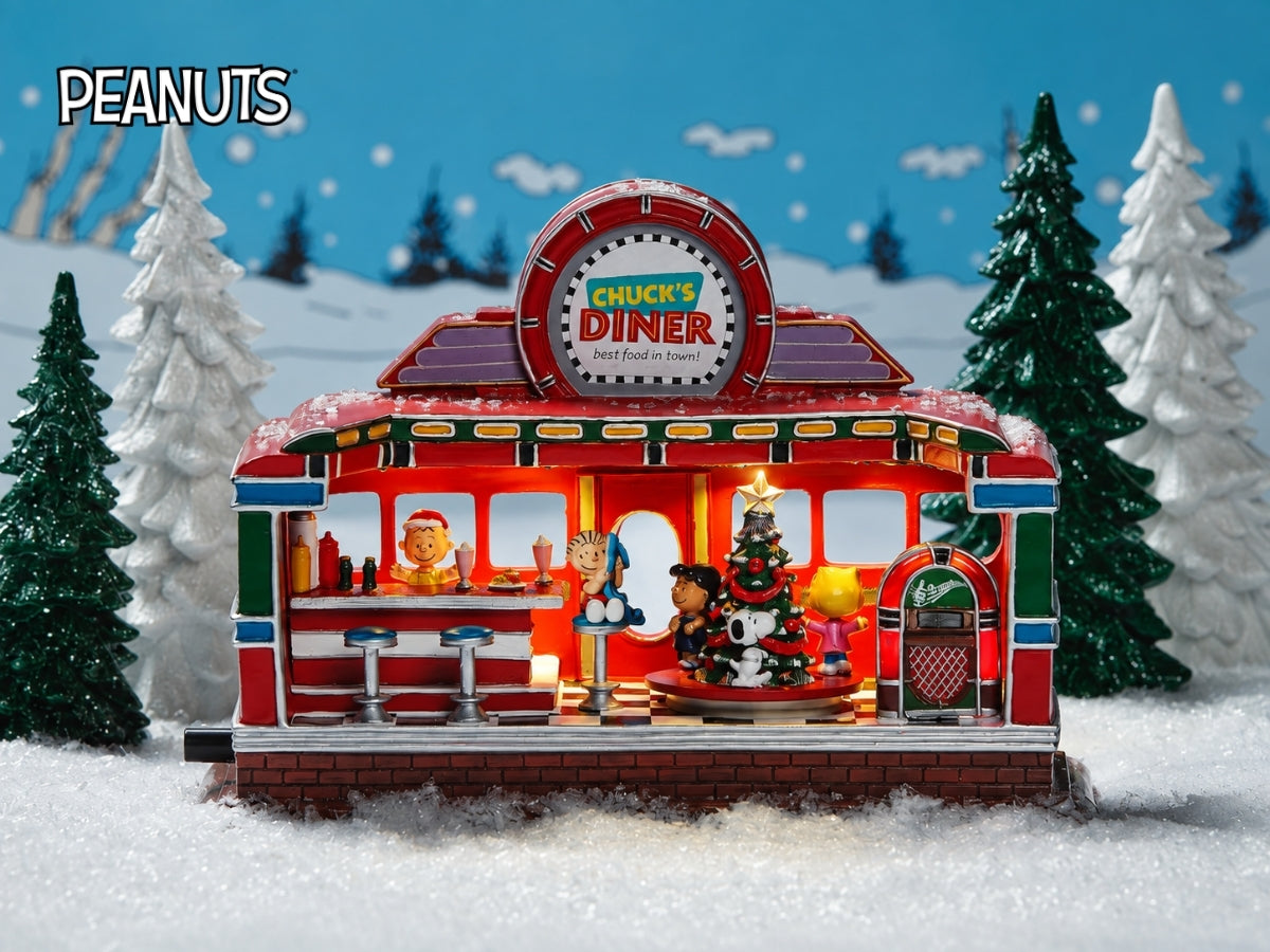 A Peanuts-themed miniature 'Chuck's Diner' set in a snowy winter scene. The red and green retro diner features a Christmas tree, small character figures, and a sign reading 'CHUCK'S DINER'.