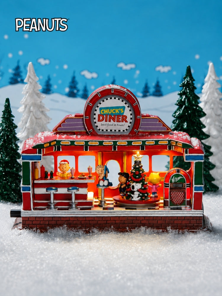 A decorative miniature diner set from the Peanuts series, featuring a red and white 'Chucky's Diner' with lit windows, a Christmas tree, and characters inside, displayed in a snowy diorama with a blue sky backdrop.