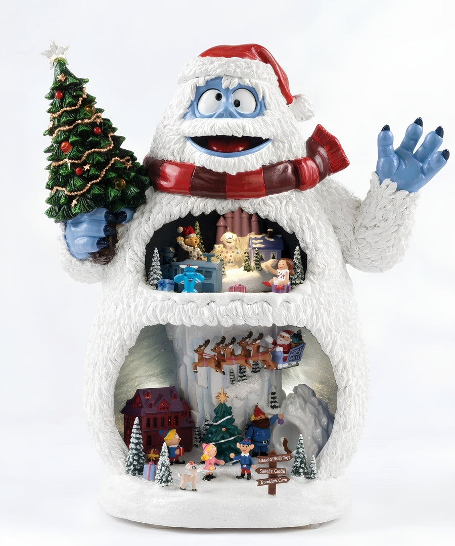 A festive white yeti figurine holding a decorated Christmas tree, with a red Santa hat and red striped scarf. The open belly area reveals a multi-level holiday scene with miniature sledding tracks, tiny houses, trees, and gift boxes, all photographed against a clean white background.