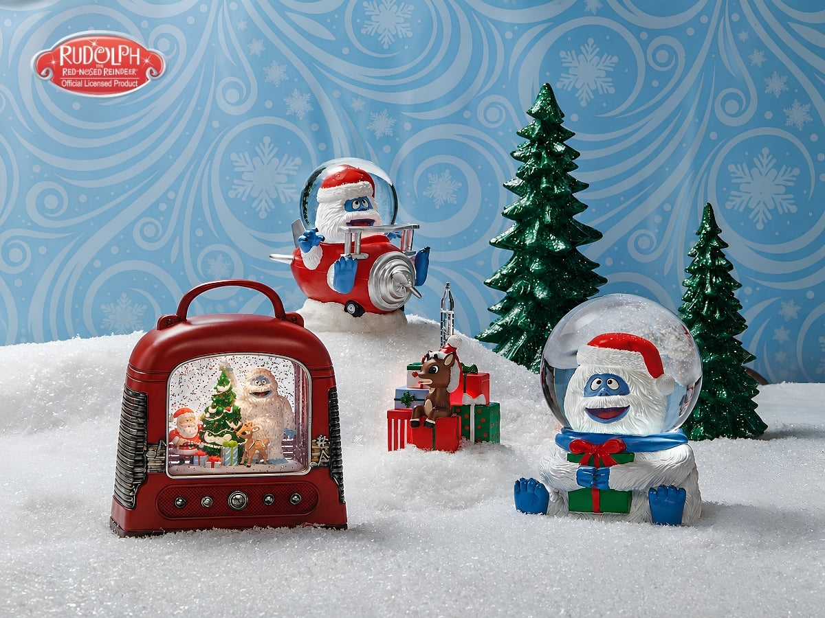 A holiday-themed product display featuring a Rudolph the Red-Nosed Christmas Santa snow globe, a red vintage radio snow globe, a snowman in a clear snow globe with a Santa hat, and a small Rudolph airplane figure, arranged on white faux snow against a blue patterned backdrop with green trees.