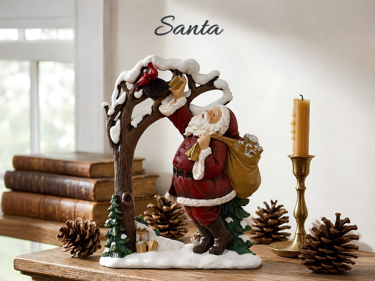A Santa Claus holiday figurine standing under a snow-dusted tree, holding a sack and reaching for a ornament, displayed on a wooden surface with books, pinecones, a candle, and the word 'Santa' above.