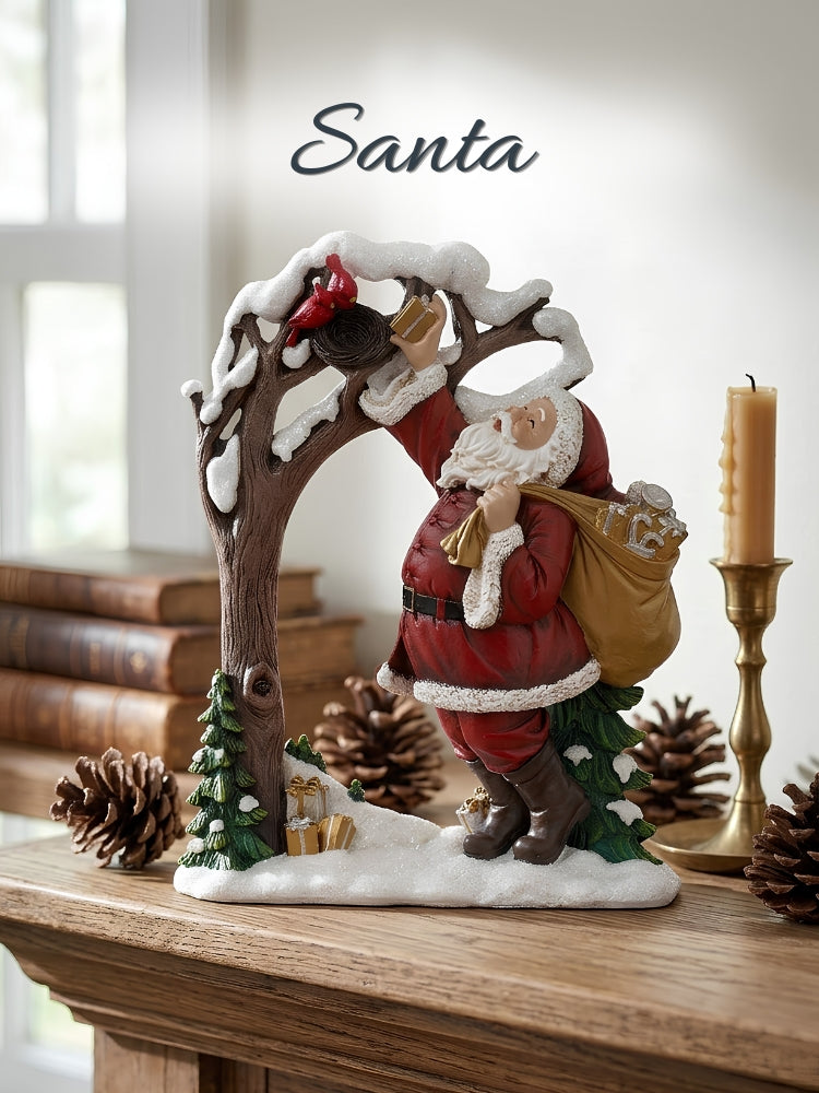 A Santa Claus holiday figurine standing beneath a snowy tree arch, with a sack on his shoulder, displayed on a wooden surface with pinecones, books, and a candle in a softly lit indoor setting. The word 'Santa' appears above the figurine.