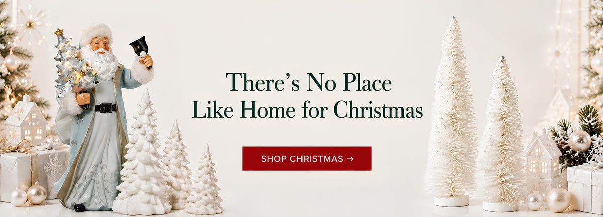 Christmas-themed promotional banner with a Santa Claus figurine holding a gift and phone, surrounded by white decorative Christmas trees and ornaments on a light background. Large text reads 'There's No Place Like Home for Christmas' with a red 'SHOP CHRISTMAS' button below.