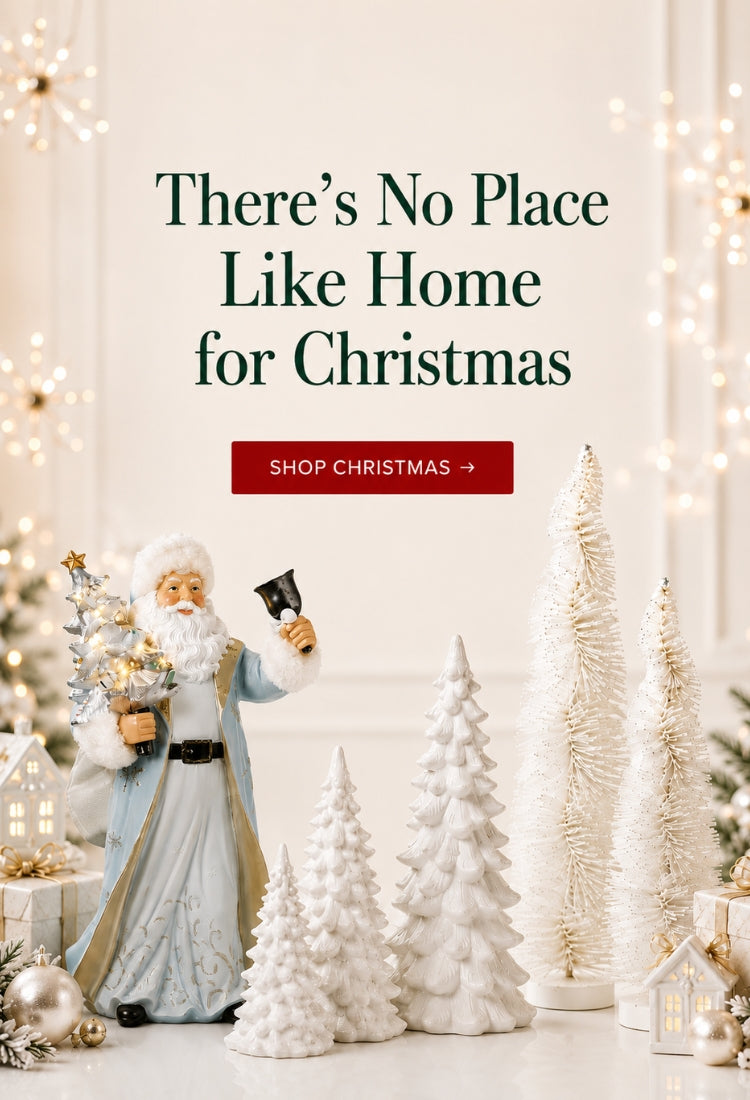 A festive holiday scene with a Santa Claus figurine holding a small decorated tree and a black bell, surrounded by white textured Christmas trees, warm string lights, and decorative lanterns. Large text reads, 'There’s No Place Like Home for Christmas' with a red button below that says 'SHOP CHRISTMAS'.